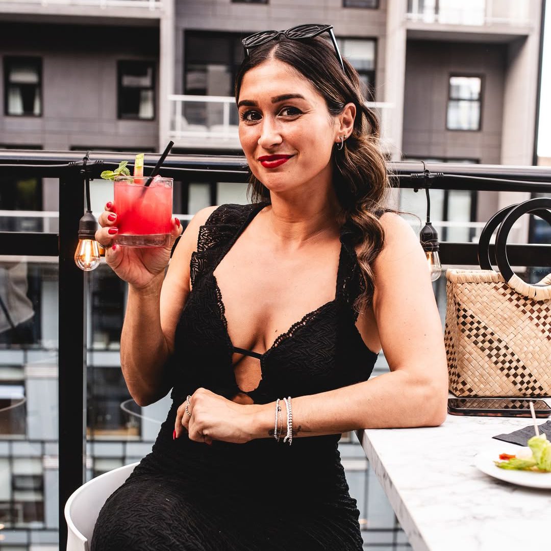 Femme élégante en robe noire savourant un cocktail sur une terrasse chic avec vue urbaine à Montréal.