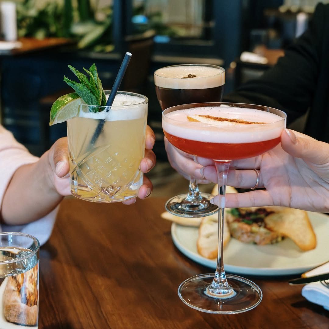 Trois cocktails artisanaux portés en toast à la Brasserie Milton, ambiance conviviale et plats gourmands en toile de fond.