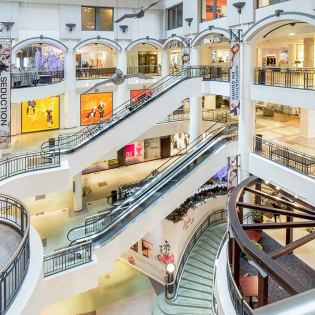  Intérieur des Cours Mont-Royal à Montréal, montrant des escaliers mécaniques, des magasins et des affiches publicitaires.