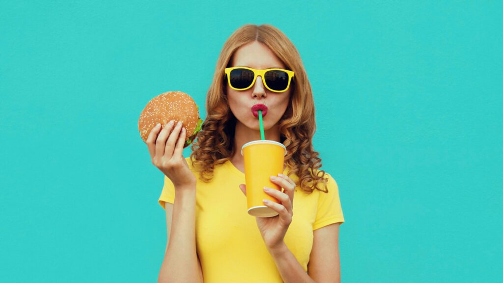 Woman in yellow sunglasses drinking a soda and holding a burger, against a bright blue background.