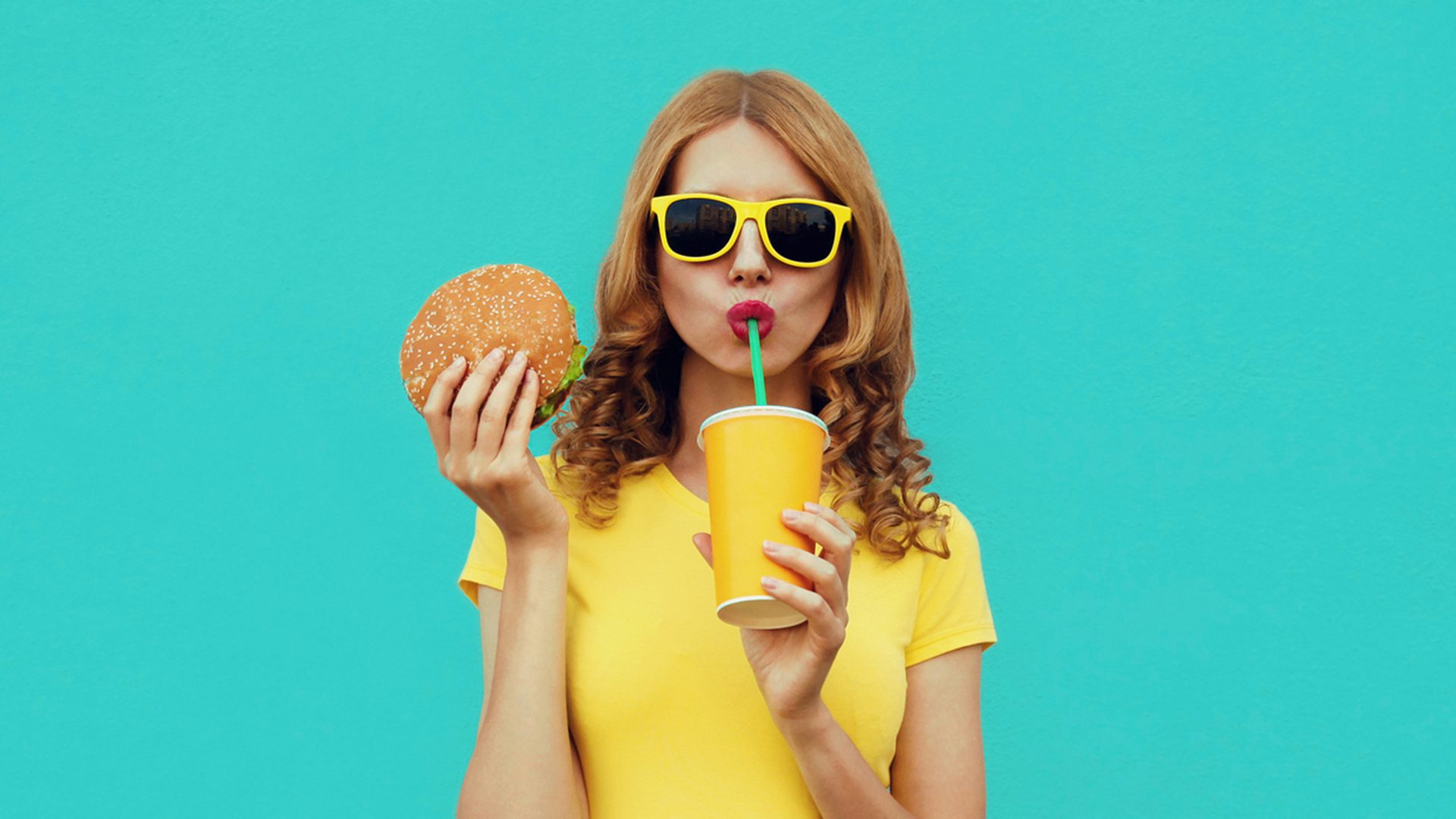 Femme en lunettes jaunes buvant un soda avec un hamburger, devant un fond bleu vif.