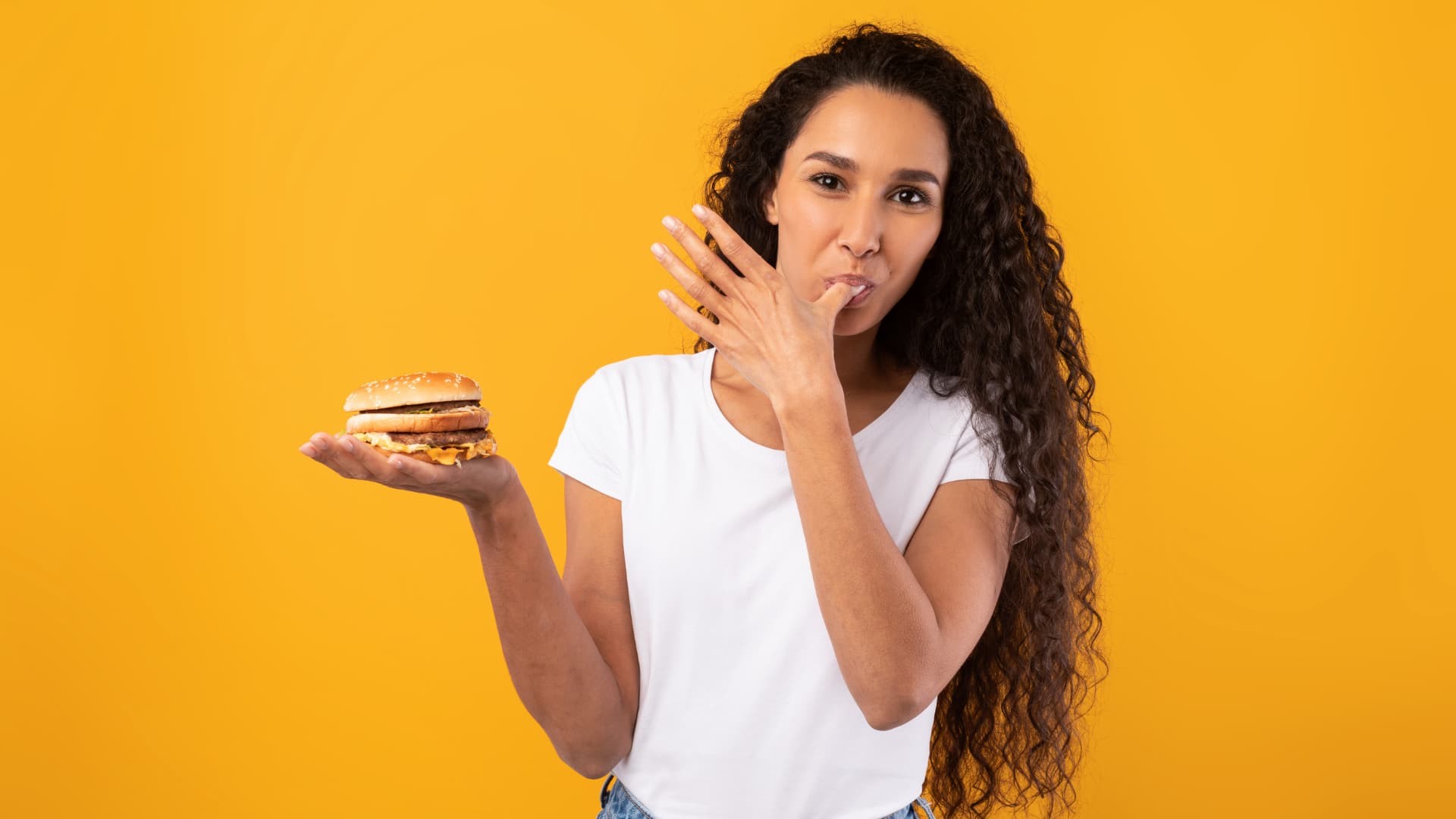 Woman in a red cap eating a burger and holding a drink.