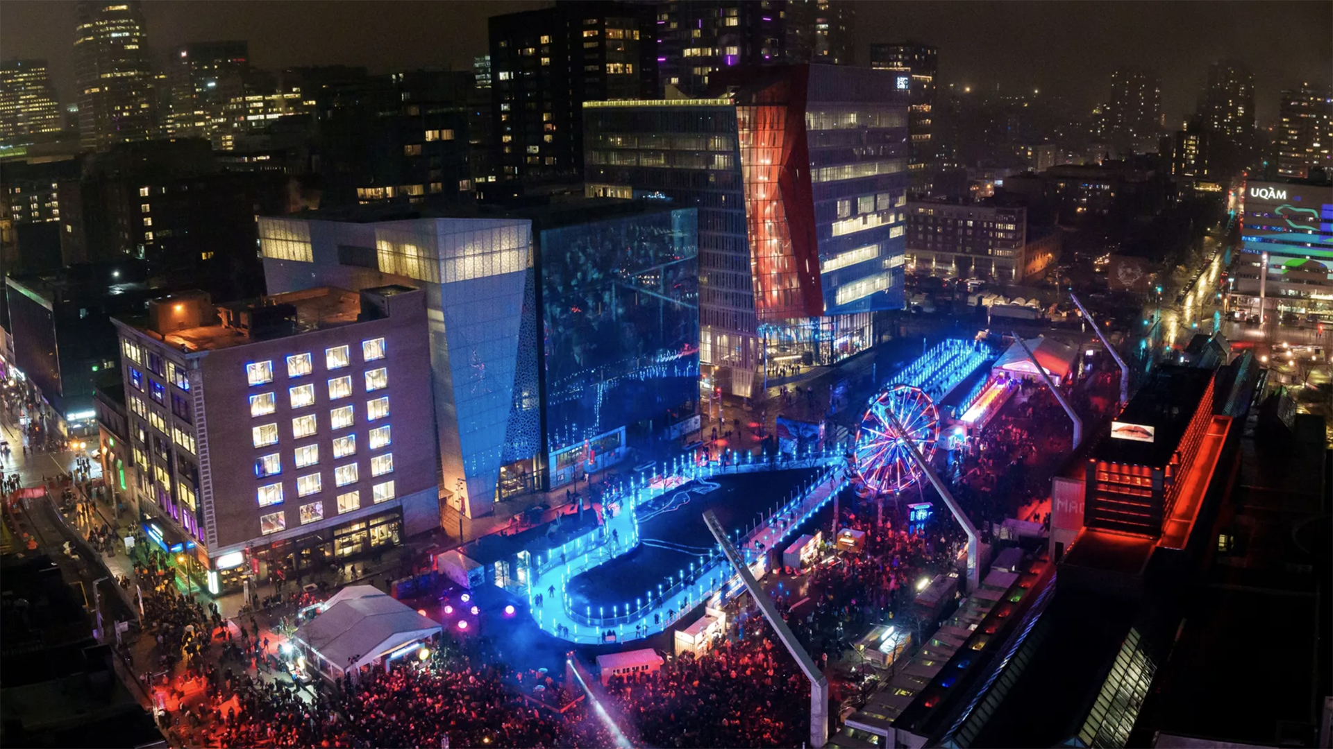 Night view of the Montréal en Lumière festival at Quartier des spectacles with a Ferris wheel, skating rink, and colorful lights.