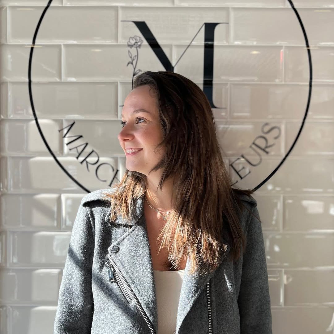 Lioudmila Zoueva smiling in front of the Marché aux Fleurs logo, illuminated by natural light.