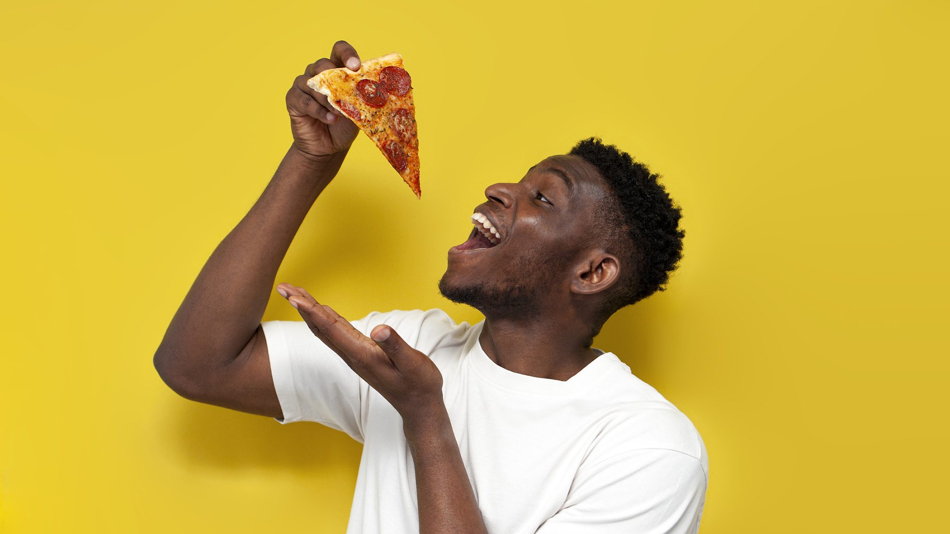 A smiling man about to take a bite of pizza against a vibrant yellow background.