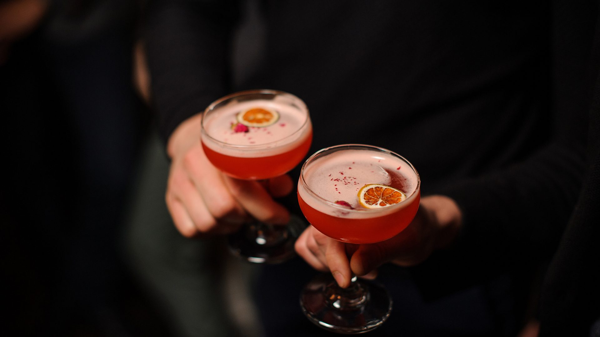 Two pink cocktails in coupe glasses, garnished with dried citrus slices, in a dimly lit setting.