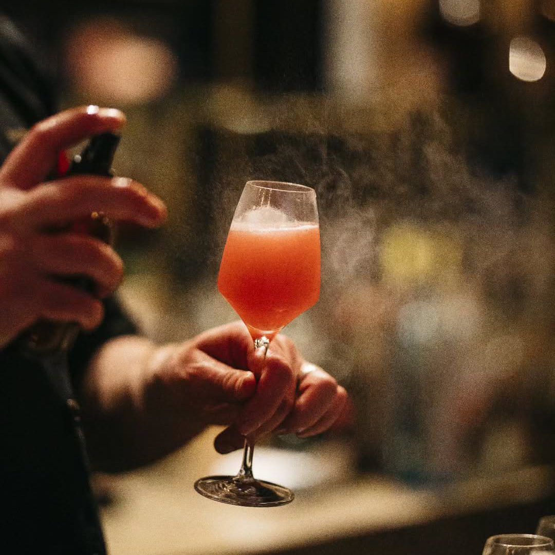 Bartender misting a pink cocktail in a stemmed glass.