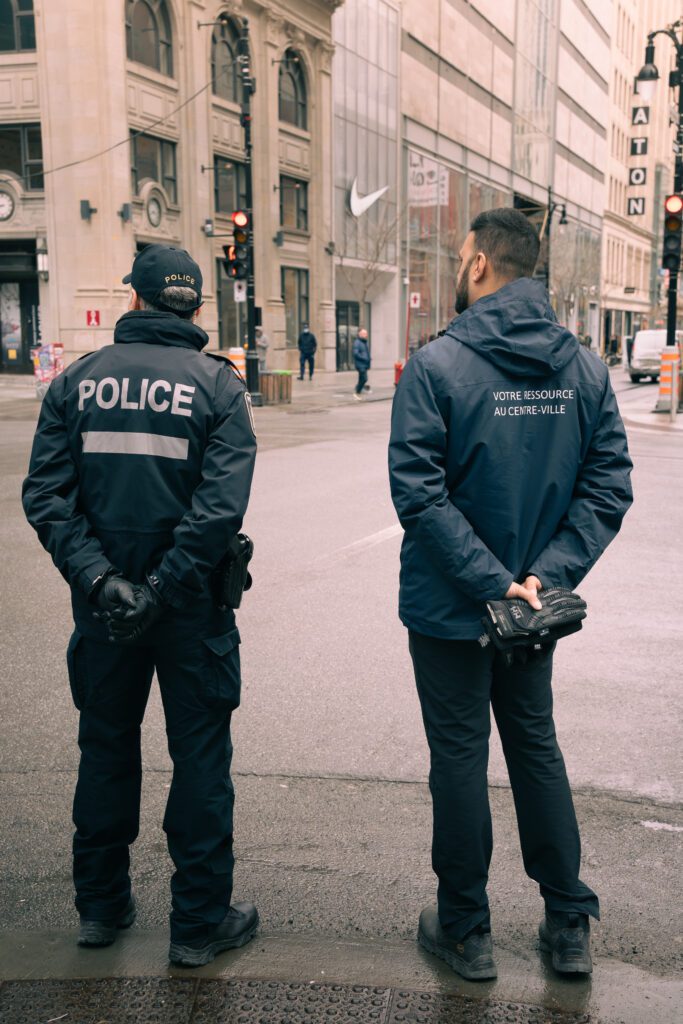 Un policier et un employé de Montréal centre-ville regardent la rue Sainte-Catherine, ils sont vus de dos. 